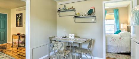 Un coin repas avec une table et des chaises, une étagère fixée au mur où sont rangées de la vaisselle, et une porte qui donne sur une autre pièce.