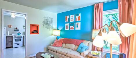 Una sala de estar con sofá, mesa de centro y lámpara. Se ve un área de cocina a través de una puerta abierta.