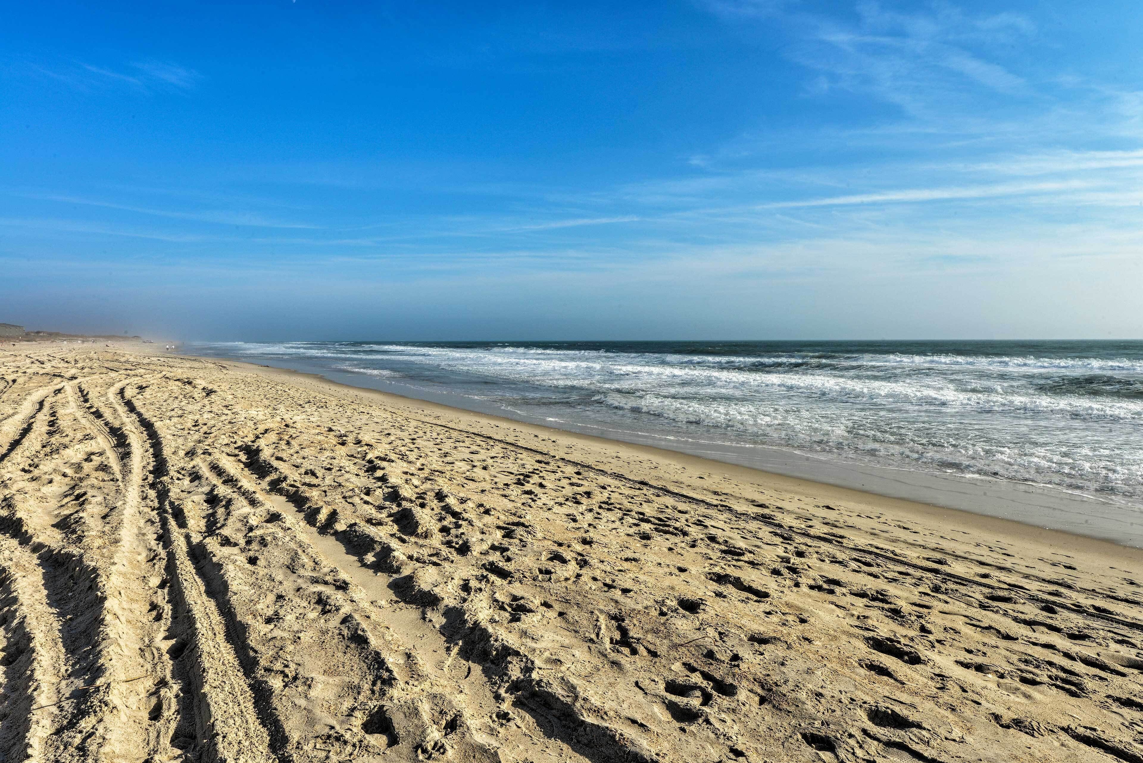 A sandy beach with tire tracks leading to the water, a clear sky, and waves approaching the shore.