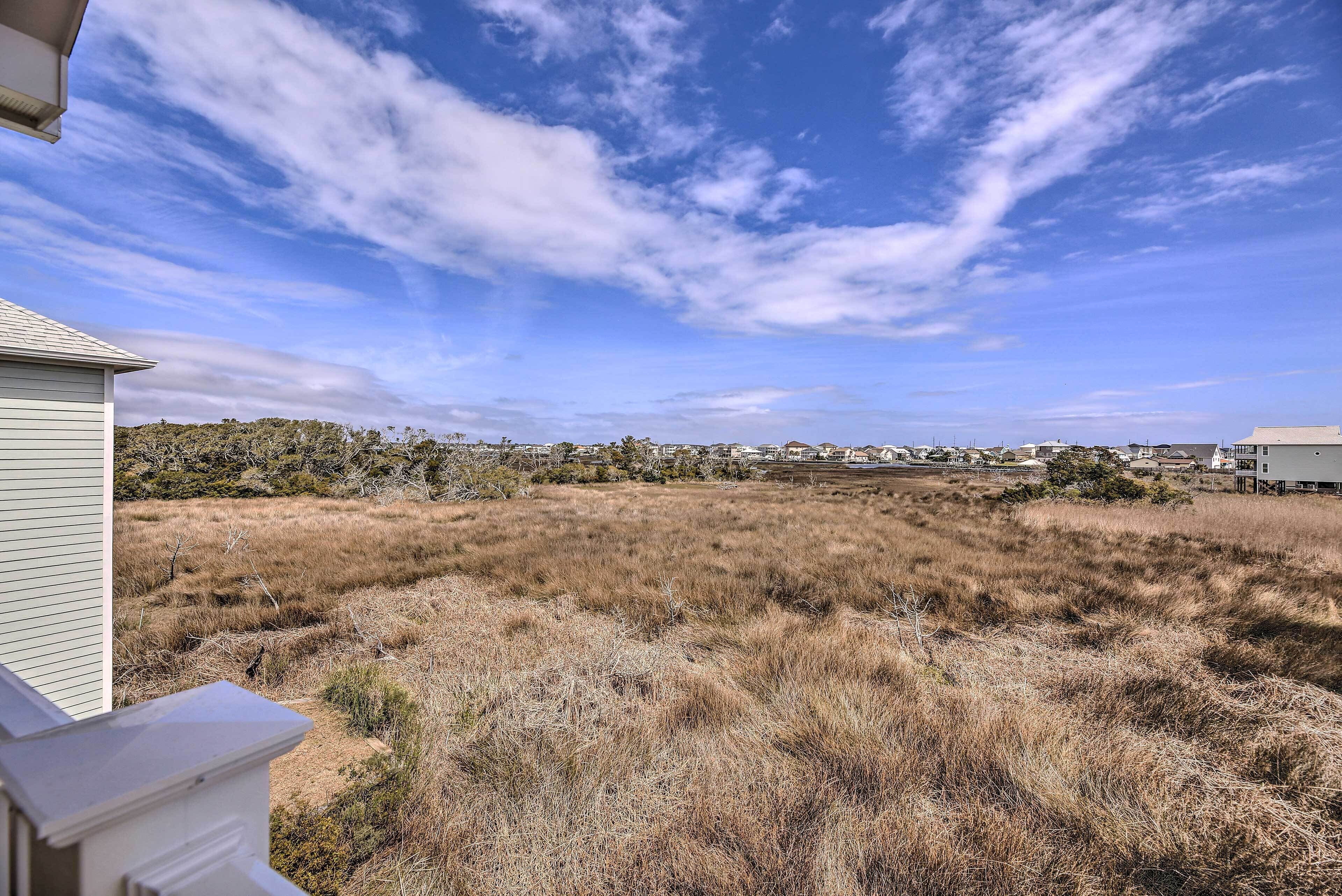 A residential area with a dry grassy field, a white building with a sloped roof, and a clear blue sky with scattered clouds.