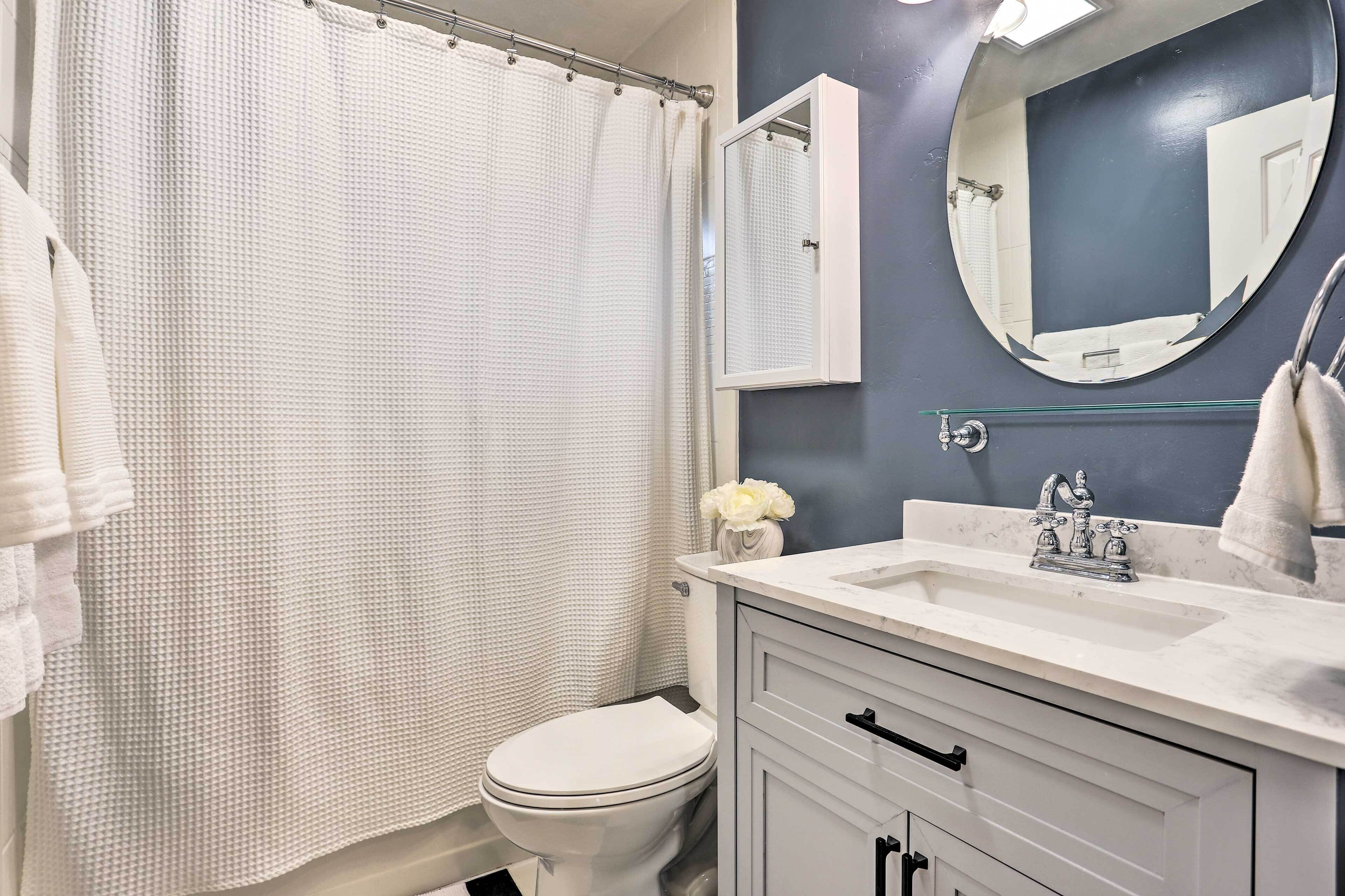 Une salle de bain avec des toilettes blanches, un lavabo blanc doté d’un plan de travail en marbre, un rideau de douche blanc et un miroir au-dessus du lavabo.