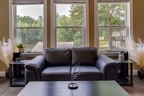A modern living room with a leather sofa, a coffee table, and a view of a tree-lined yard through large windows.