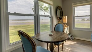 A dining area with a round table and two chairs, large windows with a view of a grassy area and a body of water, and a mirror on the wall. - Rockport Condo ~ 6 Mi to Rockport Beach Park! (Rockport)