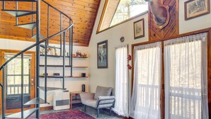 Un escalier en colimaçon mène à une chambre à coucher située en mezzanine, avec un plafond en bois, une tête de cerf accrochée au mur et un coin salon confortable comprenant un fauteuil et une petite table.
