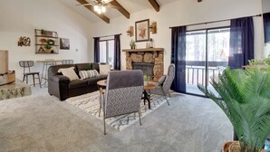 A living room with a stone fireplace, a ceiling fan, and a view of the outdoors through large windows. - Forest-view Balcony: Pinetop-lakeside Condo (Pinetop)