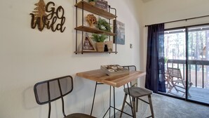 A dining area with a wooden table and chairs, a decorative wall shelf with plants, and a view of the outdoors through a sliding glass door. - Forest-view Balcony: Pinetop-lakeside Condo (Pinetop)