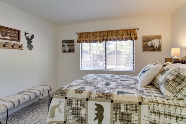 A bedroom with a bed, a bench, a window with blinds, and wall decorations including a deer head and a sign.