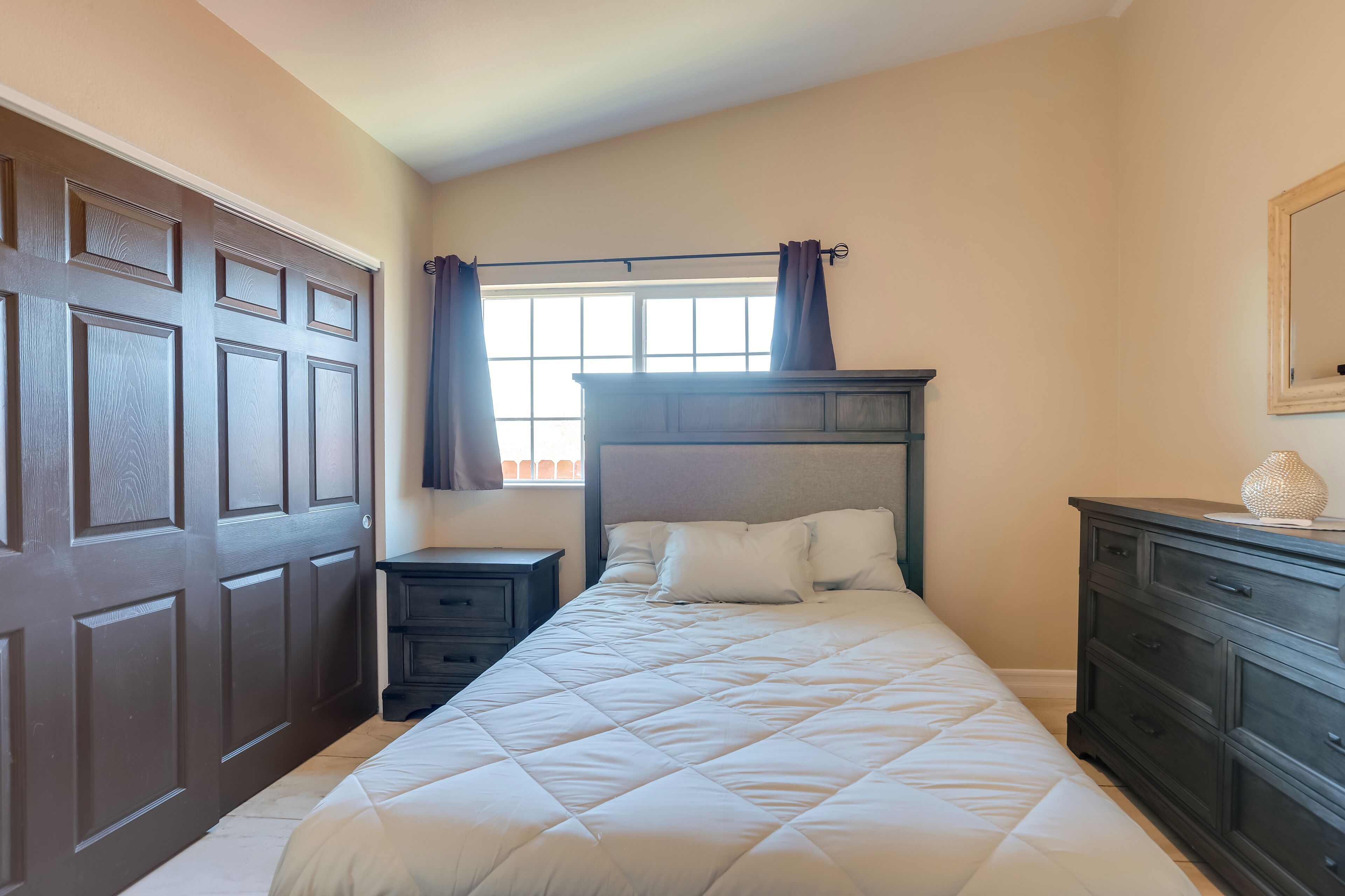 A bedroom with a large bed, a dresser, a nightstand, and a window with curtains.