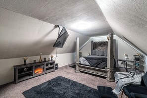 Une chambre mansardée avec une cheminée, un lit, un téléviseur fixé au mur et une petite table avec une lampe.