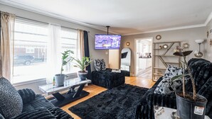 Ett vardagsrum med en platt-TV, en krukväxt, en svart soffa och ett soffbord i trä.