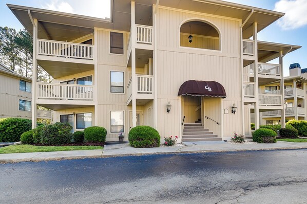 A multi-story building with balconies, a covered entrance, and a parking lot in front.