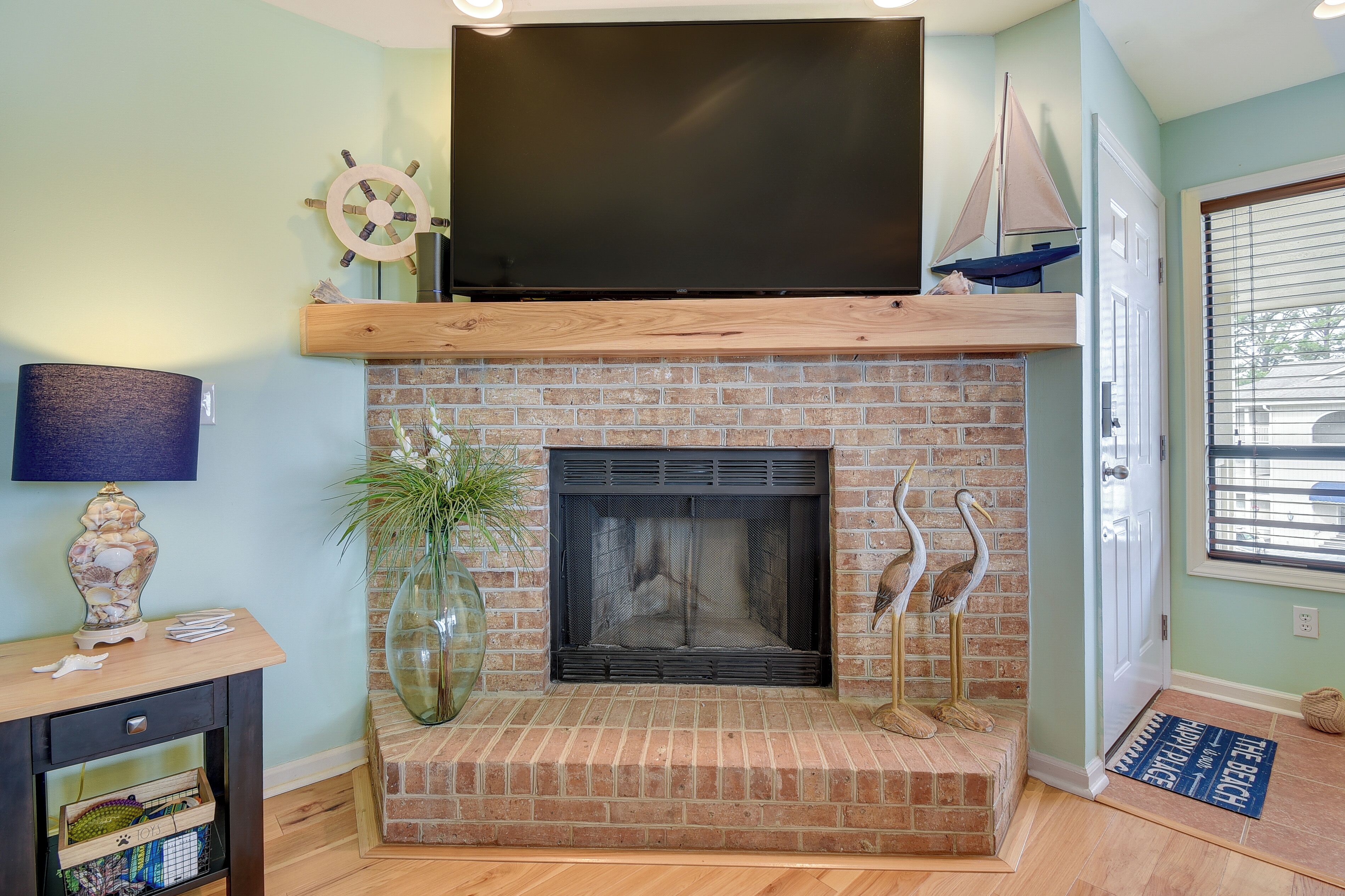 A living room with a brick fireplace, a flat-screen TV mounted above it, a wooden console table with decorative items, and a large window with blinds.