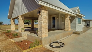 A single-story house with a covered porch supported by stone pillars, a concrete driveway, and a landscape with small plants and a tree. - Lamesa Vacation Rental w/ Covered Patio & Fire Pit (Lamesa)