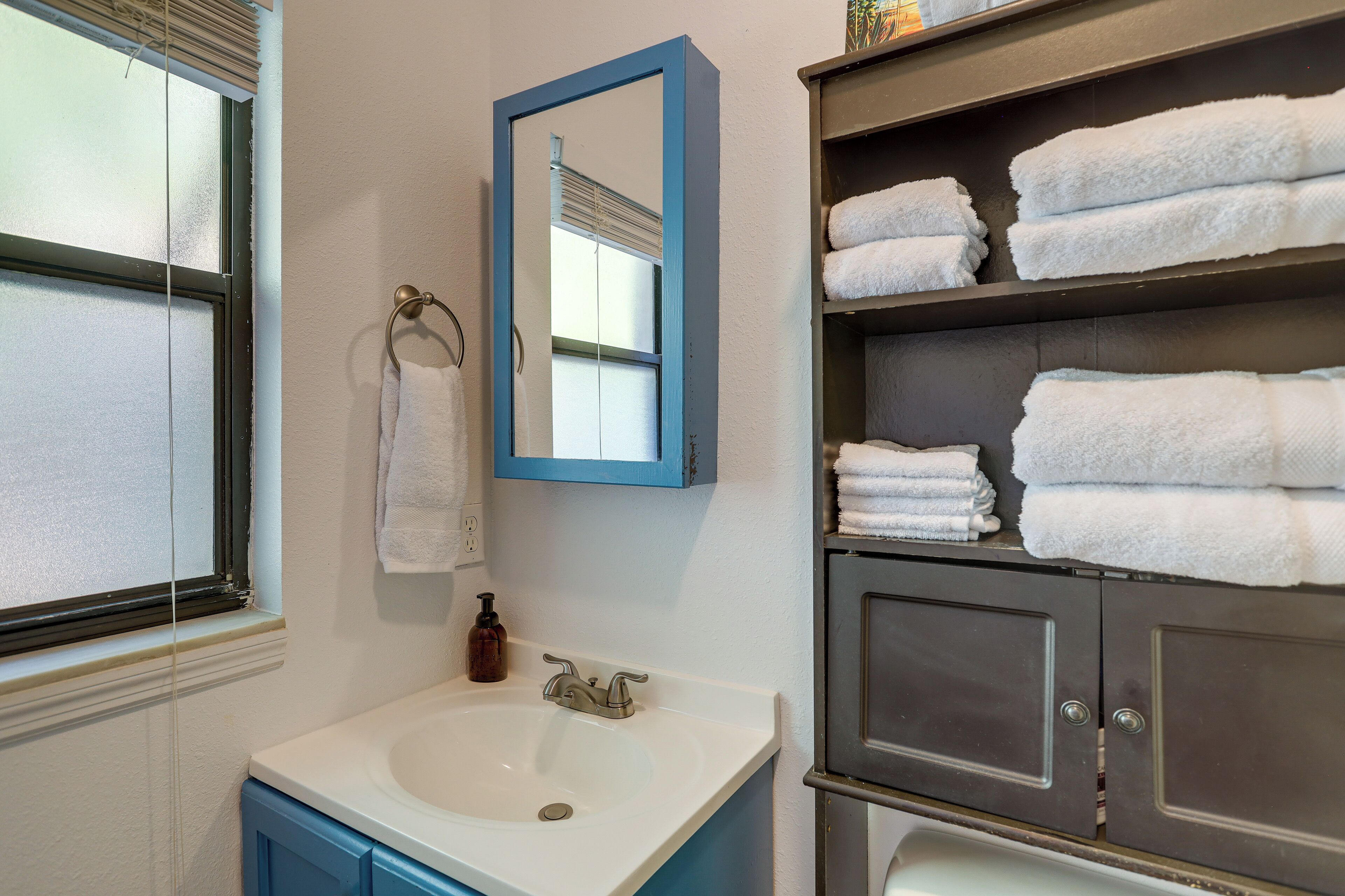 Un bagno con lavandino bianco, uno specchio con cornice blu e un porta asciugamani. Diversi asciugamani bianchi sono impilati su mensole.