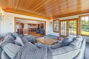 Een ruime woonkamer met een houten plafond, grijze gestoffeerde banken, een houten salontafel en uitzicht door grote ramen.