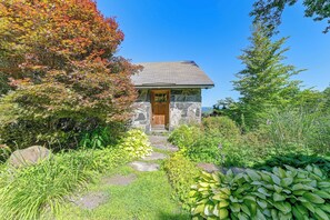 Une maison en pierre avec une porte en bois, entourée d’une végétation luxuriante et sous un ciel bleu azur.