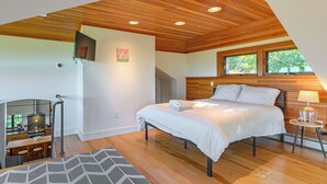 Une chambre avec un plafond en bois, un lit recouvert de draps blancs, une table de chevet avec une lampe et une fenêtre donnant sur des arbres.