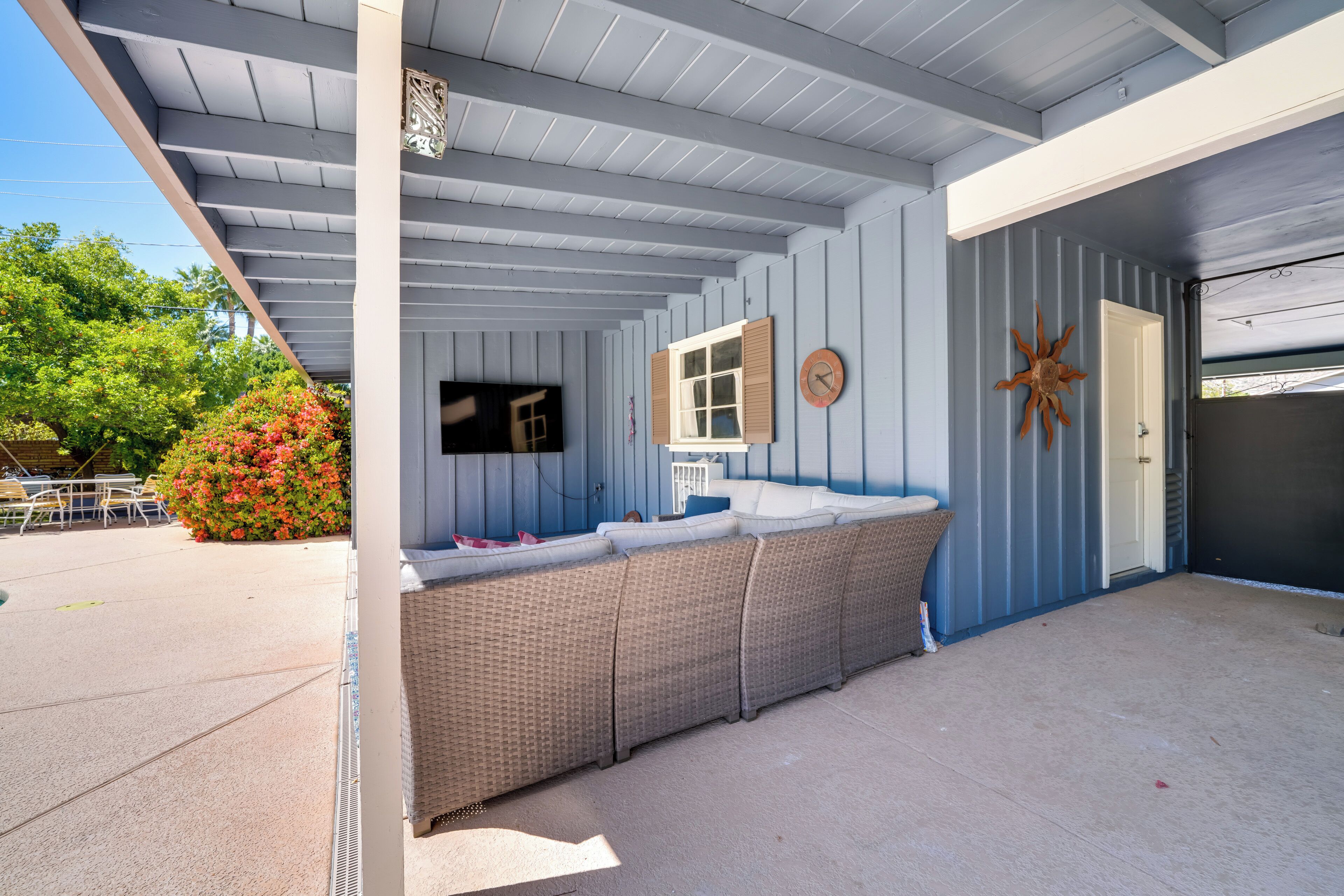 A covered outdoor seating area with a wicker sofa, a television, and a wooden door.
