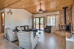 A spacious living room with a stone fireplace, a ceiling fan, and a large window. - Cozy Pine Mountain Cabin: Hike, Bike, Fish Nearby! (Pine Mountain Club)