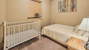 Una camera da letto con una culla, un letto, una finestra con tende, una ventola e un quadro appeso al muro.