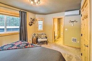 Ein Schlafzimmer mit einem Bett, einem Sessel, einem Fenster mit Vorhängen, einem Ventilator und einer Tür zu einem anderen Raum.