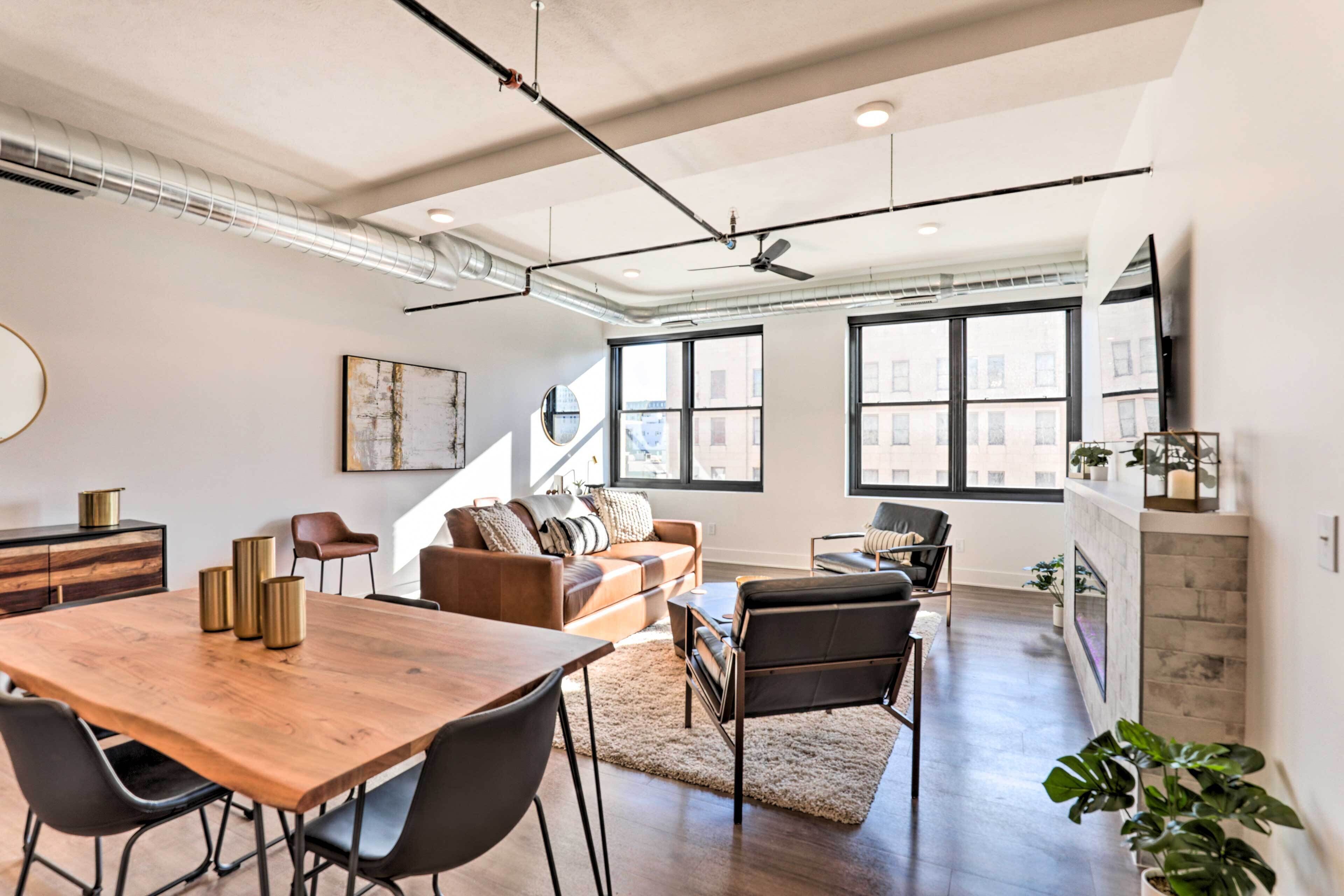A spacious living area with a wooden dining table, black chairs, a brown leather sofa, and a black leather chair. There are large windows with a view of buildings outside.
