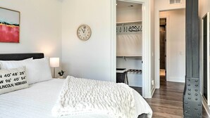 Een slaapkamer met een groot bed, een wekkerklok op het nachtkastje, een klok aan de muur en een kledingkast met daarin hangende kleding.