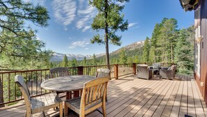 Eine Holzterrasse mit rundem Tisch und Stühlen, umgeben von einer Brüstung, mit Blick auf eine gebirgige Landschaft.