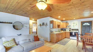 A cozy living room with a sofa, coffee table, and a dining area with chairs and a table. A kitchen with wooden cabinets and appliances is visible in the background. - Cozy Heber Cabin Retreat w/ Deck + Fireplace! (Overgaard)