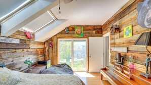 Ein Schlafzimmer mit schrägverdeckter Decke, Holzwänden, einem Bett mit gemusterten Betttextilien, einer Bank und einer Tür, die nach draußen führt.