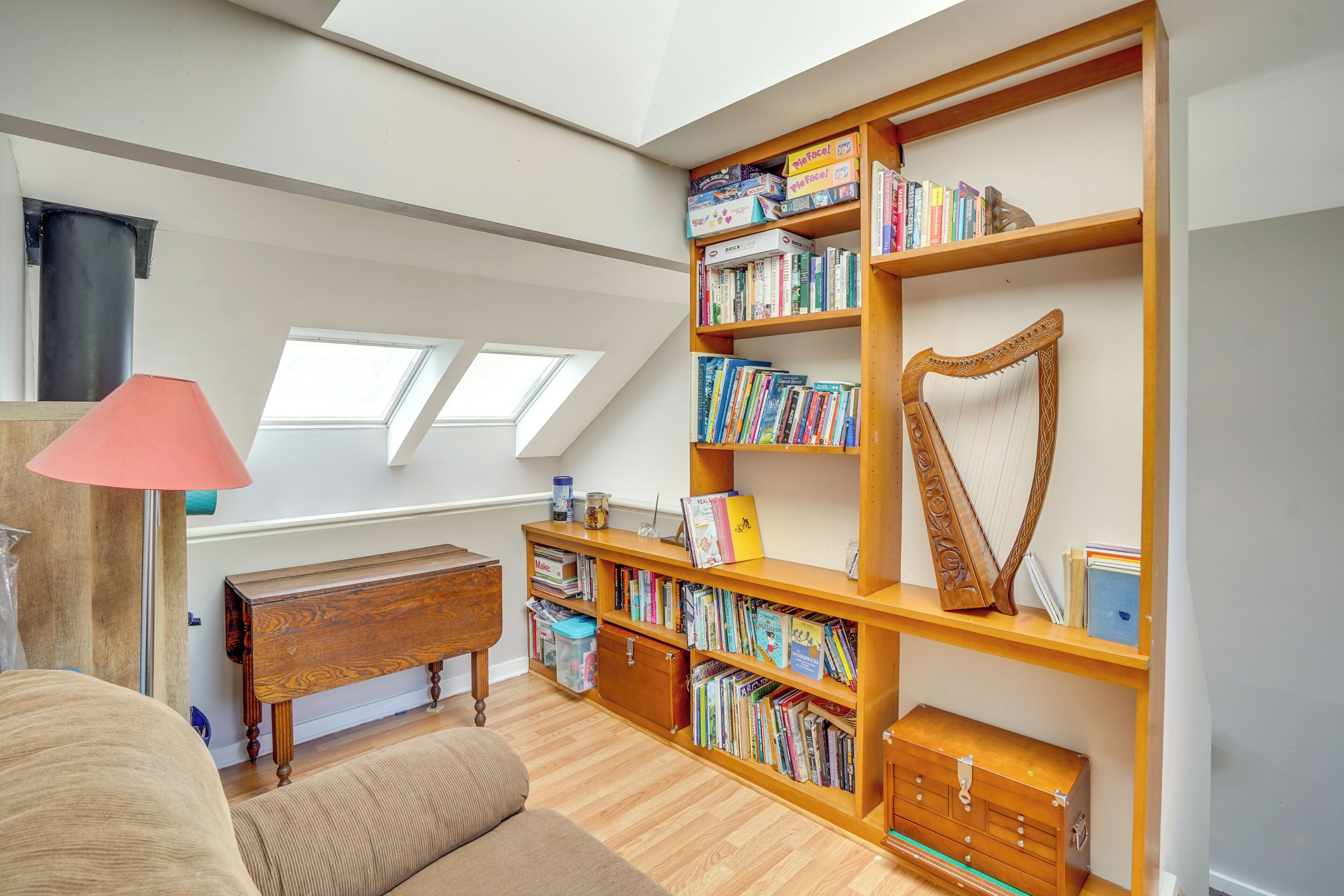 A spacious loft bedroom with a large skylight, a wooden bookshelf filled with books, a harp, and a cozy seating area.