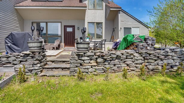 Ein zweistöckiges Haus mit einer Steinmauer davor, einer Grillkota und einer Terrassenfläche.