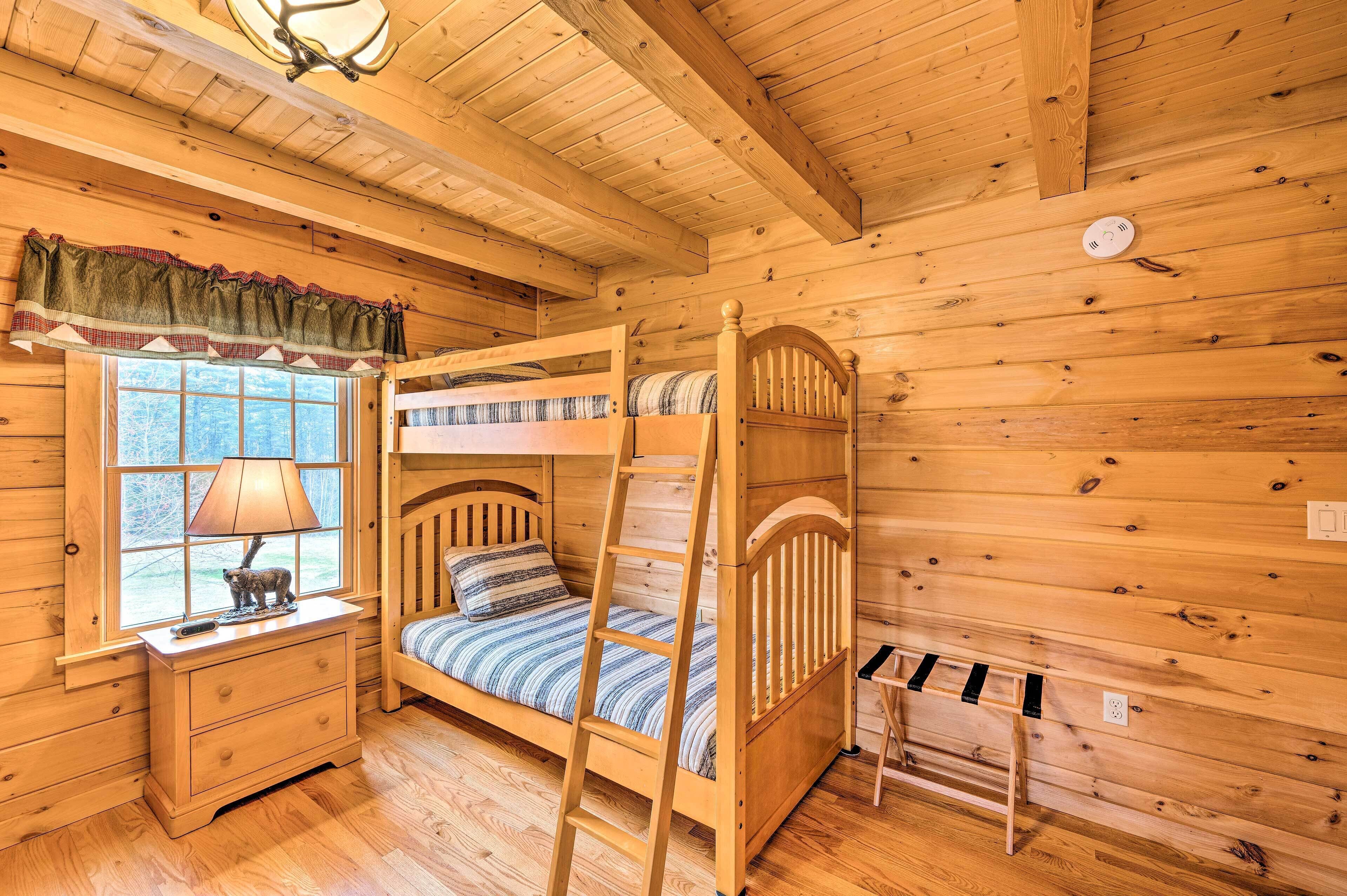 A wooden cabin room with a bunk bed, a nightstand with a lamp, and a window with a view of trees.