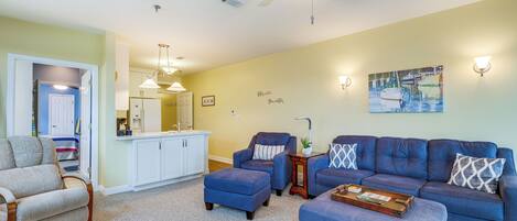 A living room with a blue sofa, matching ottoman, and a grey armchair. There is a ceiling fan, a painting of boats, and a kitchen area in the background.