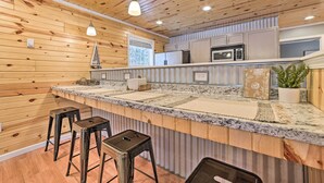 A modern kitchen with wooden paneling, granite countertops, stainless steel appliances, and bar stools. - Cozy Elmira Cabin ~ 23 Mi to Boyne Mountain Resort (Mancelona)