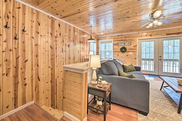 A cozy cabin interior with wooden walls, a gray sofa, a small table with a lamp, and a ceiling fan. - Cozy Elmira Cabin ~ 23 Mi to Boyne Mountain Resort (Mancelona)