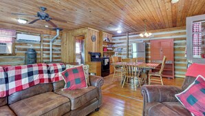 A log cabin interior with a wooden ceiling, a dining area with a table and chairs, a kitchen with appliances, and a leather sofa with a plaid blanket. - Historic Log Cabin w/ Porch Near Patoka Lake! (Taswell)