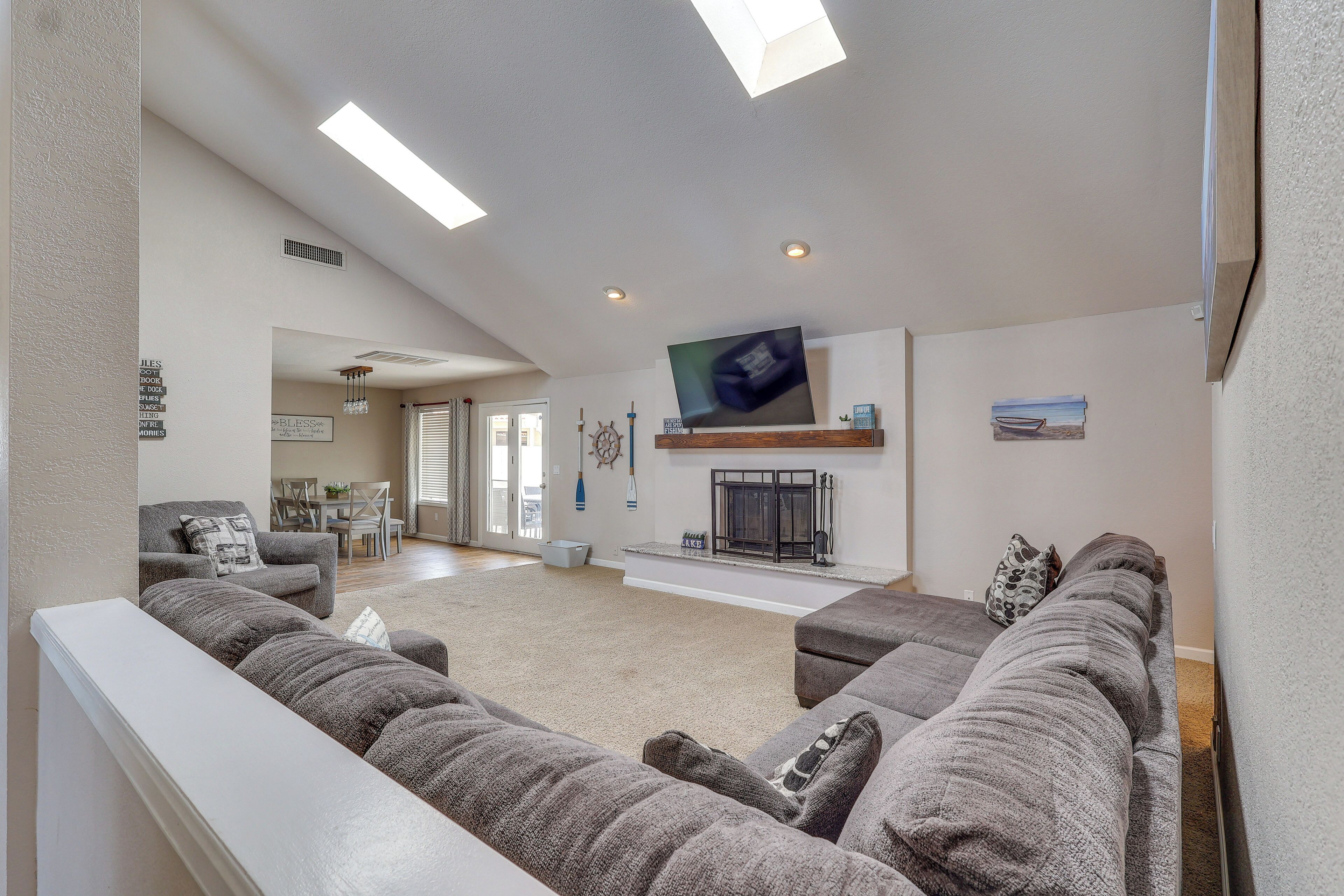 A living room with a large sectional sofa, a flat-screen TV mounted on the wall, a fireplace, and a dining area in the background.