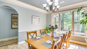 A dining room with a wooden table set for six, surrounded by chairs, a chandelier, and a large window with a view of trees. - < 5 Mi to Lake Desoto: Grand Hot Springs Home (Hot Springs Village)