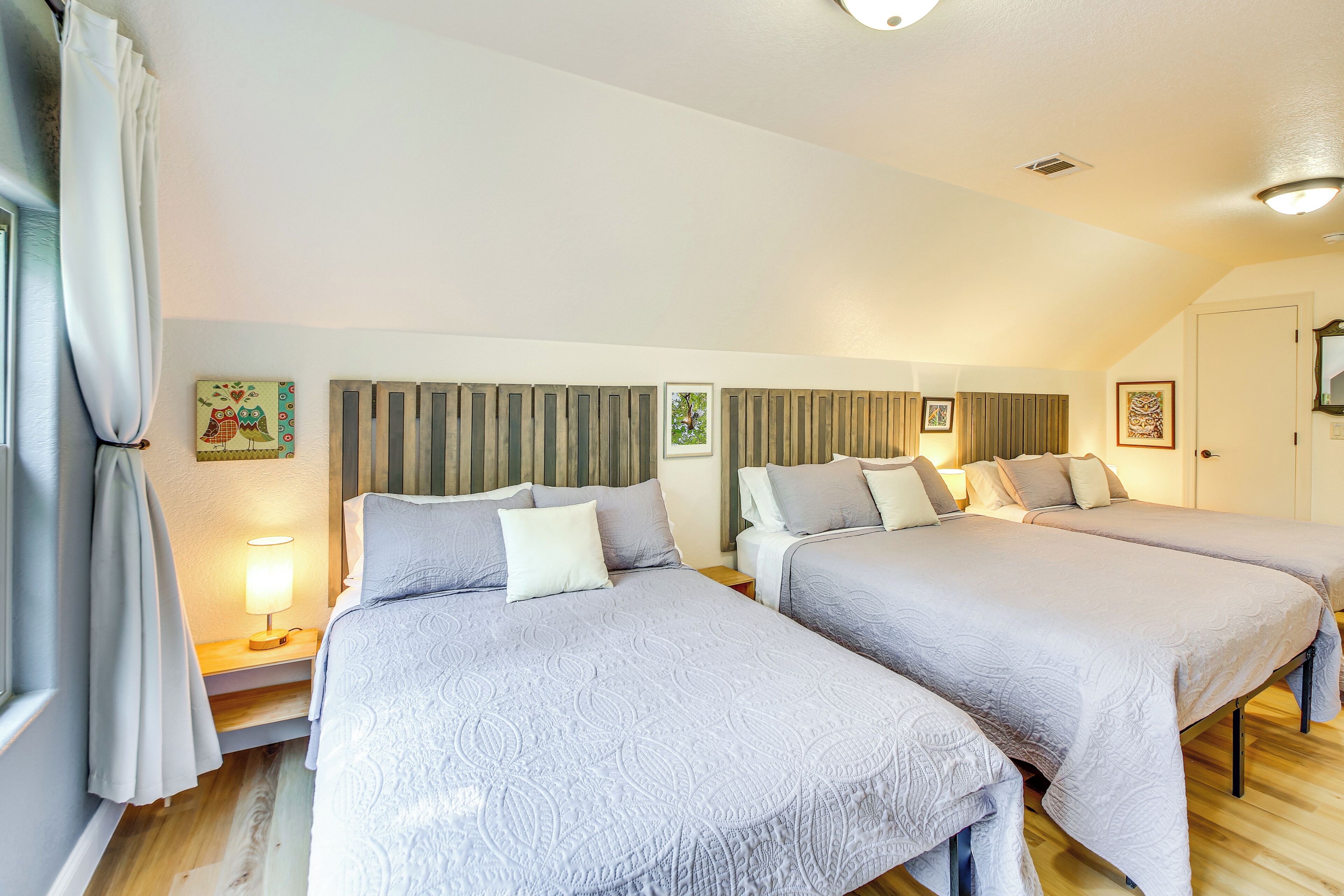 Una habitación de hotel con dos camas, una ventana con cortinas, una lámpara y obras de arte en las paredes.