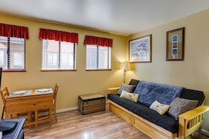 A living room with a wooden table, a black sofa, and a red-trimmed window. - Sunriver Vacation Rental w/ Community Hot Tub! (Bend)