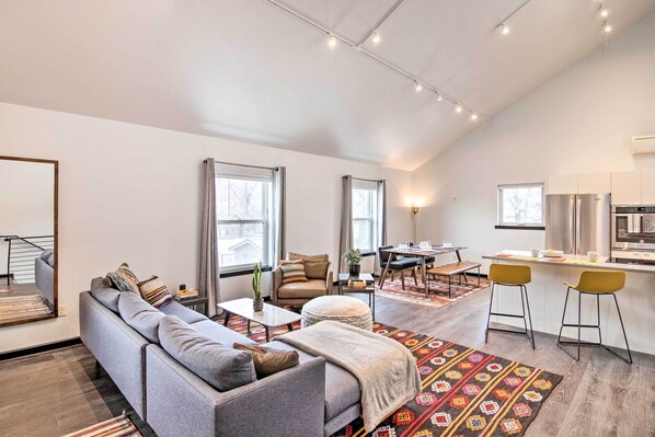 Un salon moderne avec un canapé gris, une table basse et un tapis à motifs. On y trouve également un coin repas avec une table et des chaises, ainsi qu'une cuisine équipée d'un réfrigérateur et d'un four à micro-ondes.