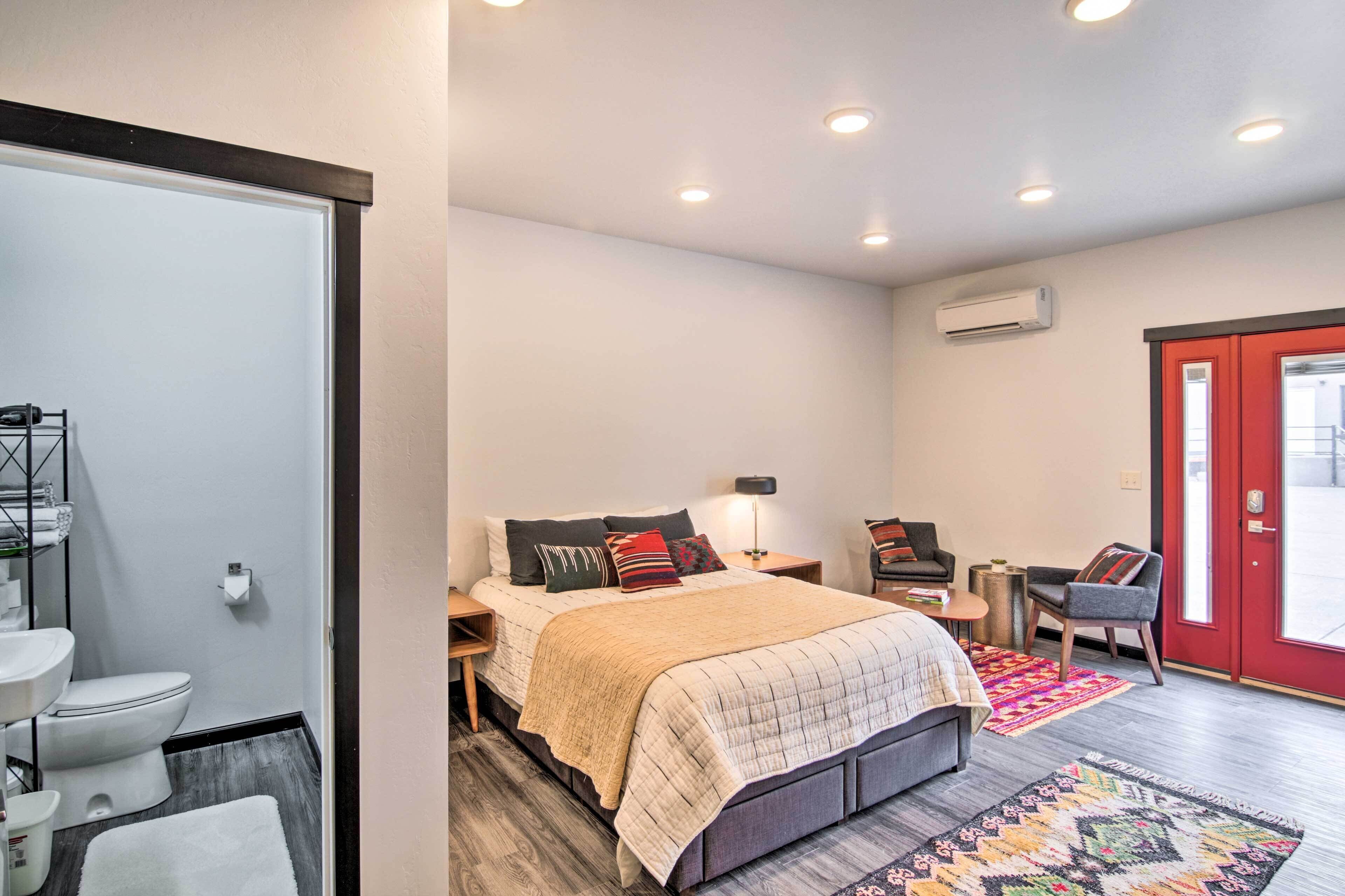 Ein Schlafzimmer mit Bett, Nachttischen, Schreibtisch, Stuhl, einer roten Tür und einem Badezimmer mit Toilette und Waschbecken.