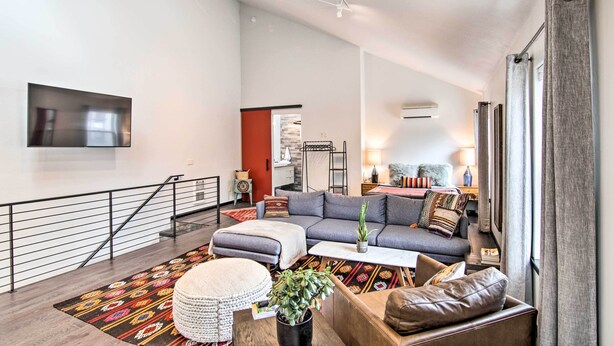 A modern living room with a grey sofa, ottoman, and coffee table, a staircase with a railing, and a flat-screen TV mounted on the wall. - Contemporary-boho Loft in Downtown Livingston! (Livingston)