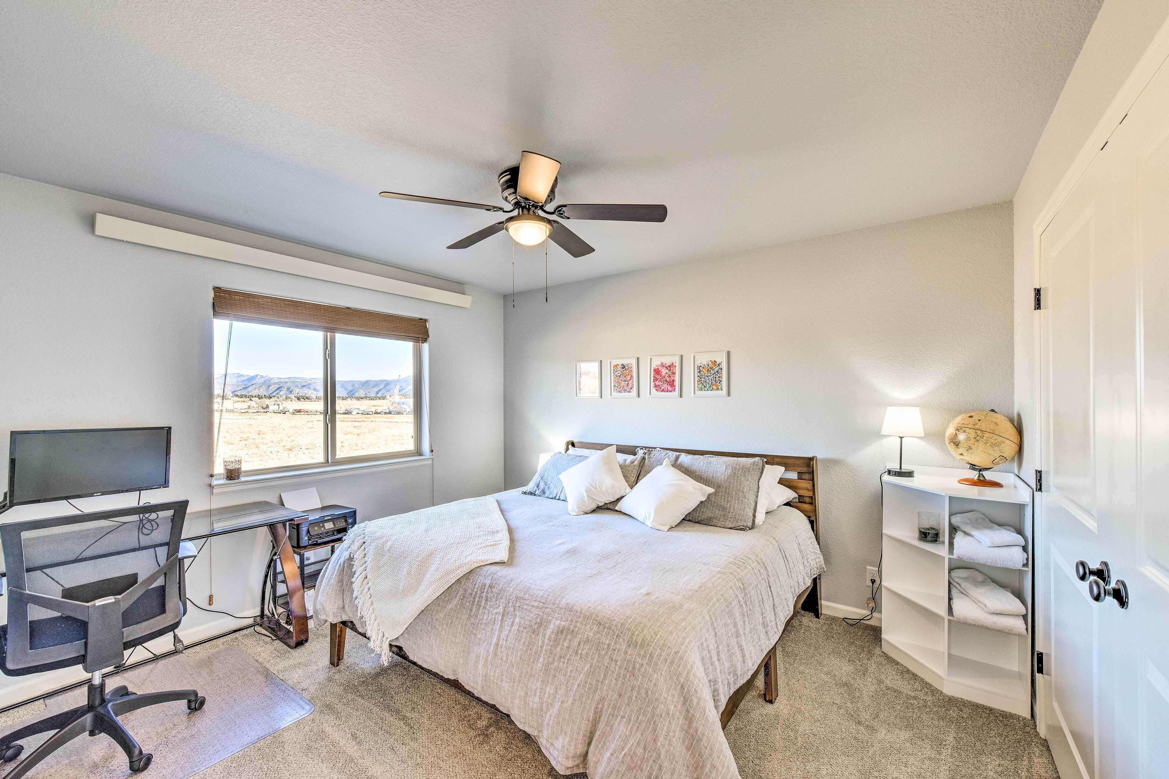 Une chambre à coucher avec un lit, un bureau avec un ordinateur, un ventilateur de plafond et une fenêtre donnant sur un beau paysage.
