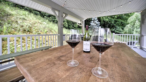 Two wine glasses on a wooden table with a bottle of wine in the background, under a covered area with a railing and greenery in the background. - Settlers Mountain Retreat: On-site Hikes & Views! (Bryson City)