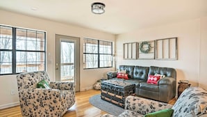 A living room with a wooden floor, a leather sofa, a patterned armchair, and a large window with a view of trees. - Renovated Cabin w/ Decks, Views, & Fire Pit! (Pikeville)