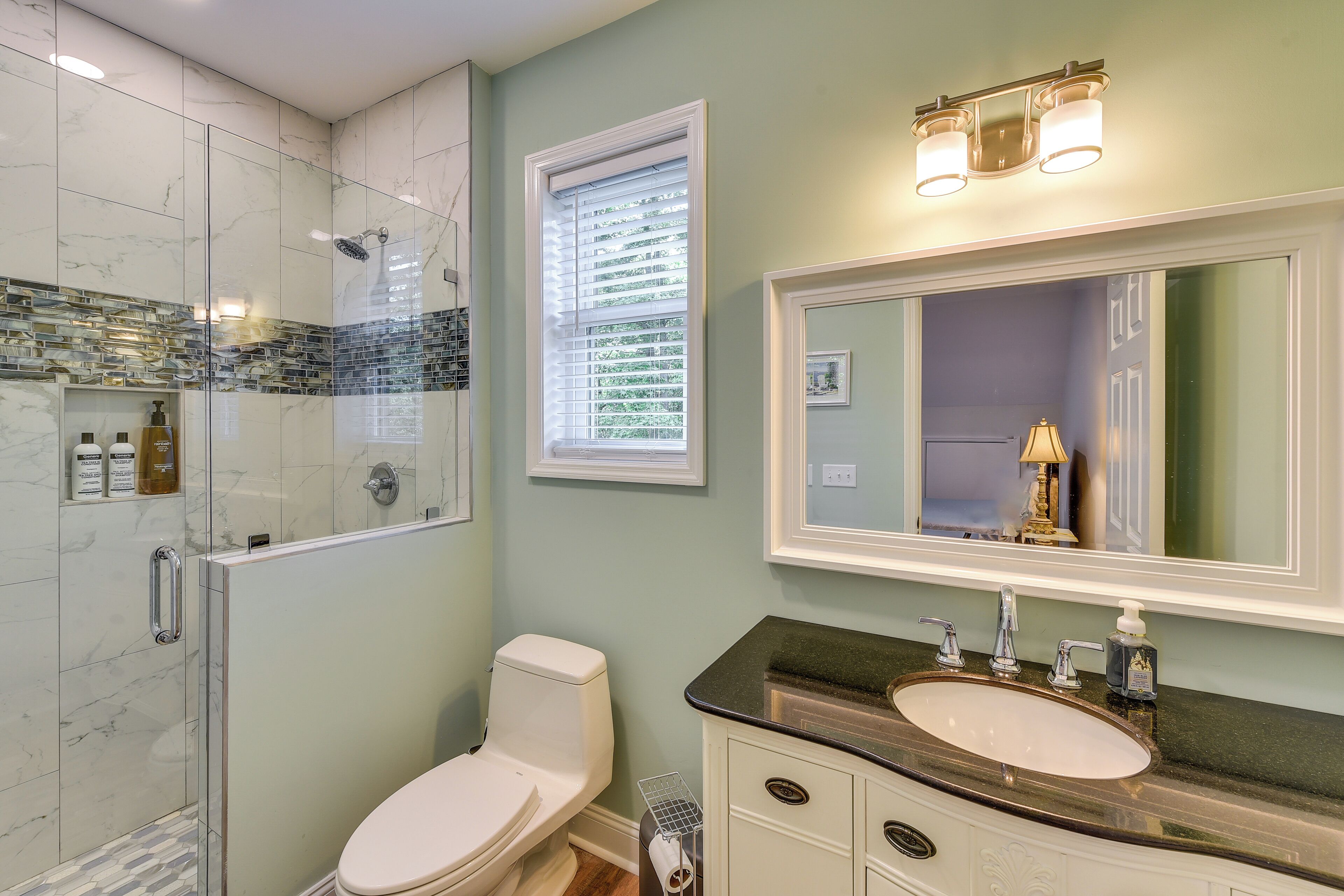 Une salle de bain avec une douche à parois de verre, des toilettes blanches, un meuble-lavabo avec un plan de travail noir et un grand miroir.