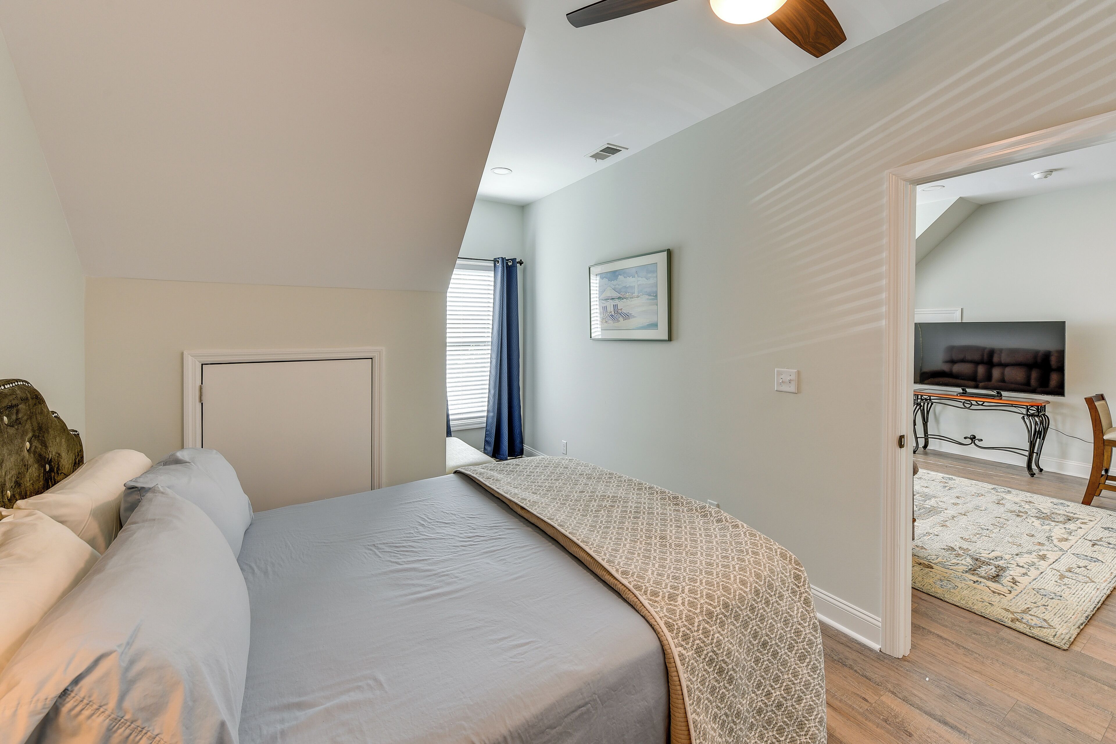 Ein Schlafzimmer mit Bett, einer Tür, einem Fenster mit Vorhängen, einem Deckenventilator und einem Fernseher im angrenzenden Raum.