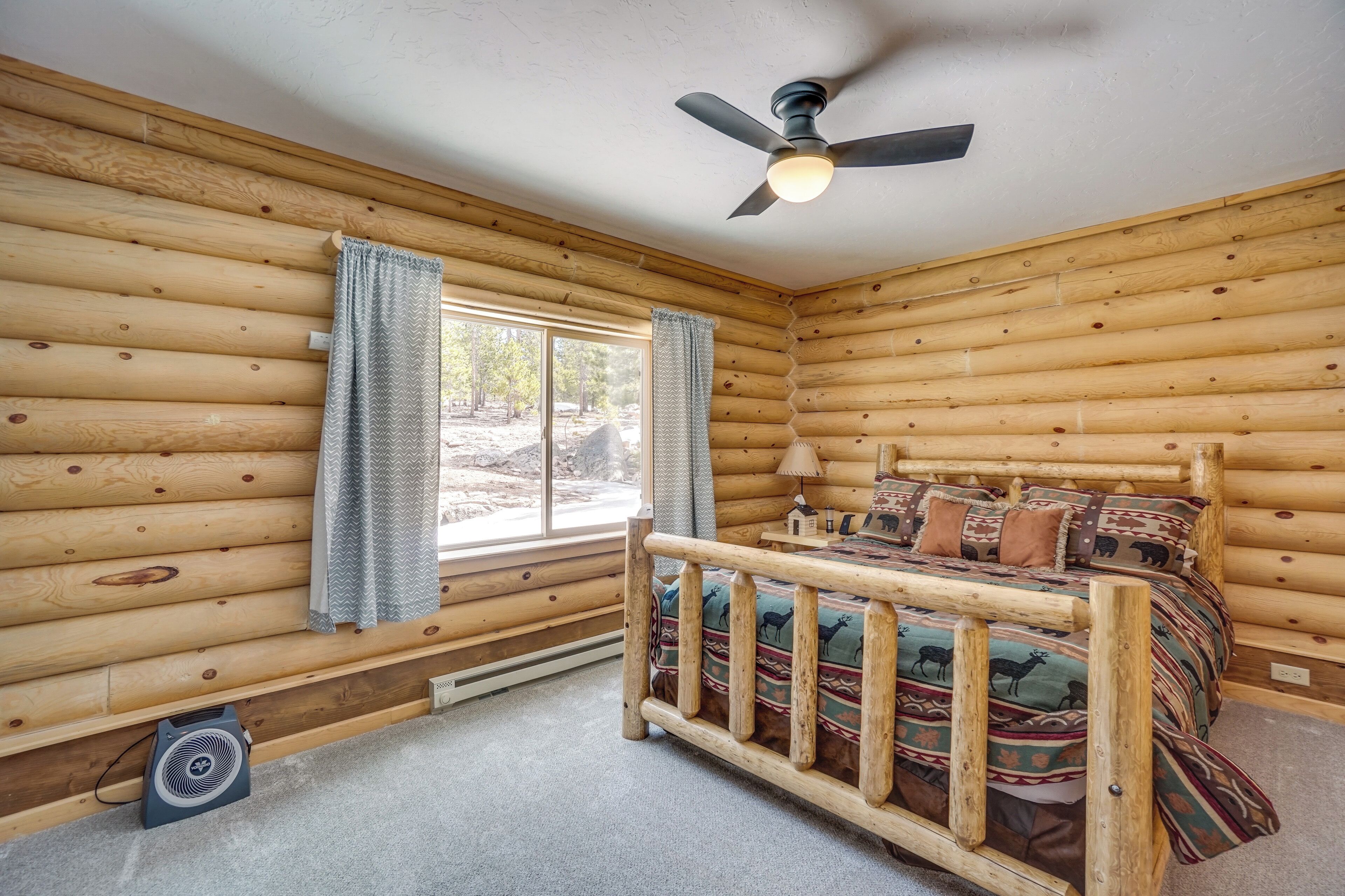 A log cabin bedroom with a bed, a ceiling fan, and a window with curtains.